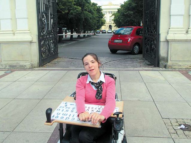 Agnieszka Bal siedzi na wózku wyposażonym w tablicę z literami. Za nią brama Uniwersytetu Warszawskiego