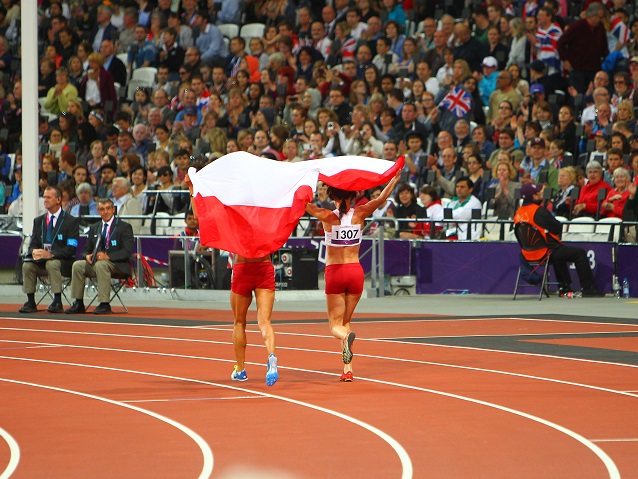 Dwie polskie zawodniczki biegną bieżnią na stadionie trzymając polską flagę