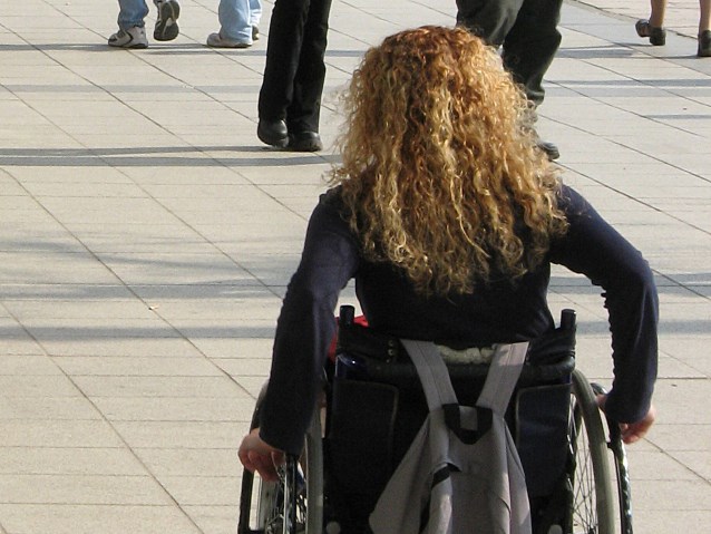 zdjęcie, przedstawiające tyłem poruszającą się na wózku kobietę z blond kręconymi włosami. na wózku z tyłu ma zawieszony siwy plecak