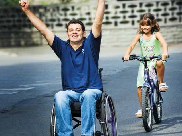 mężczyzna na wózku wyciąga ręce w geście zwycięstwa, za nim jedzie dziewczynka na rowerze