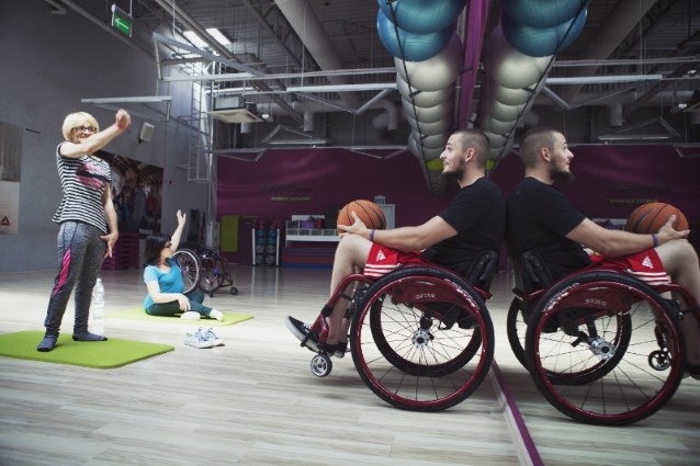 chłopak na wózku rzuca kobiecie piłkę do koszykówki, niedaleko nich ćwiczy młoda dziewczyna, przy której stoi wózek