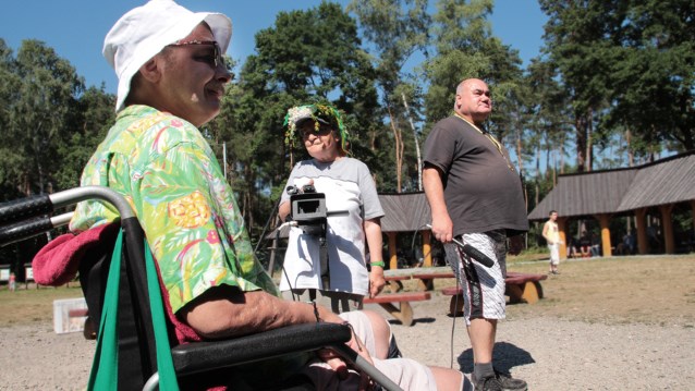 mężczyzna na wózku, obok którego są dwie osoby. Kobieta trzyma kamerę i go nagrywa