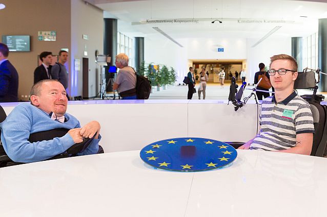 Marek Plura i Artur Wachowicz siedzą przy stole z flagą Unii Europejskiej, w tle ludzie na korytarzu