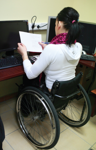Kobieta na wózku trzyma dokumenty przy biurku z dwoma komputerami