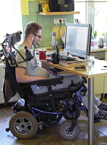 Młody mężczyzna na wózku elektrycznym pracuje na komputerze, trzymając w ustach wskaźnik