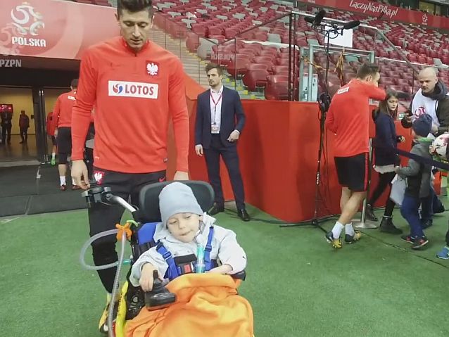 Robert Lewandowski idzie po murawie boiska za małym Frankiem poruszającym się na wózku elektrycznym