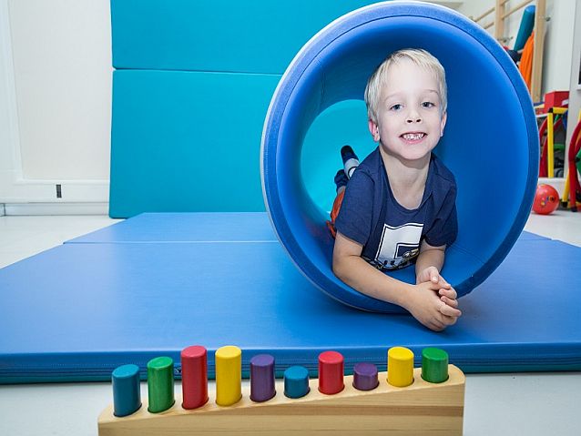 Sala ćwiczeń. Chłopiec leży w niebieskiej miękkiej tubie na materacu, z przodu zabawki edukacyjne