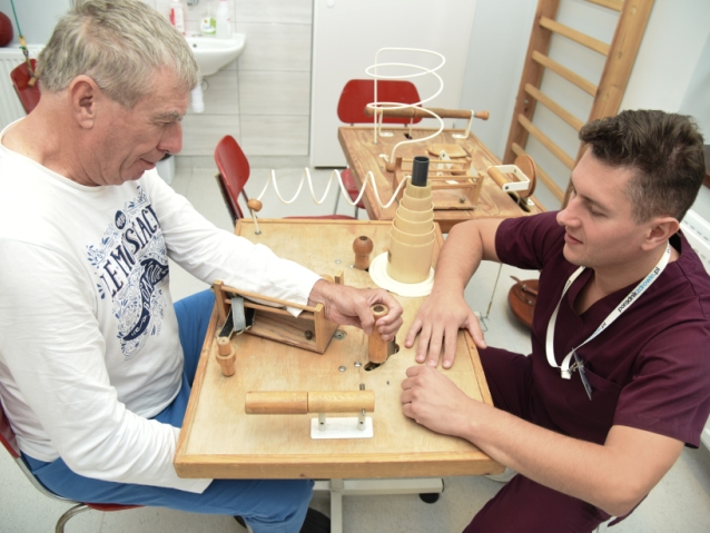 Starszy mężczyzna rehabilituje rękę i dłoń, przesuwając drążek na drewnianym urządzeniu rehabilitacyjnym, naprzeciw niego siedzi młody rehabilitant