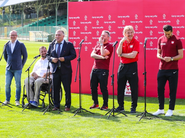 Na murawie stadionu odbywa się briefing prasowy, przy ściance Łączy nas piłka znajduje się sześć osób, przy których stoją mikrofony. Od lewej: Mariusz Trzeciakiewicz, prezes Fundacji Katarynka; Paweł Parus, członek zarządu Federacji Kibiców Niepełnosprawnych; moderator wydarzenia Dariusz Łapiński z PZPN; selekcjoner reprezentacji Jerzy Brzęczek, bramkarz Łukasz Fabiański oraz Zbigniew Boniek, prezes Polskiego Związku Piłki Nożnej