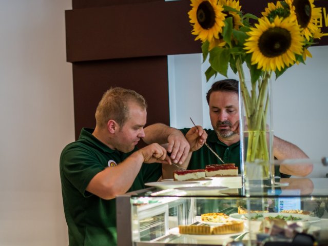 Dwóch pracowników nakłada ciasta na talerzyki, na ladzie stoją słoneczniki