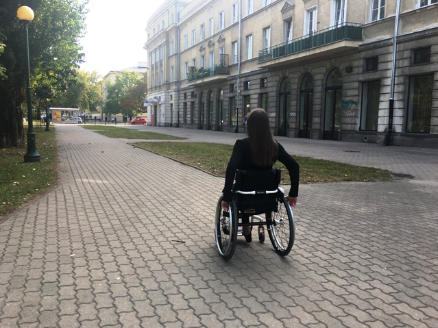 dziewczyna na wózku porusza się szerokim chodnikiem. Po prawej kamienica, po lewej drzewa i park