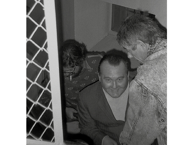 Czarno-białe zdjęcie. Dwóch mężczyzn wnosi po schodach uśmiechniętego mężczynę w średnim wieku