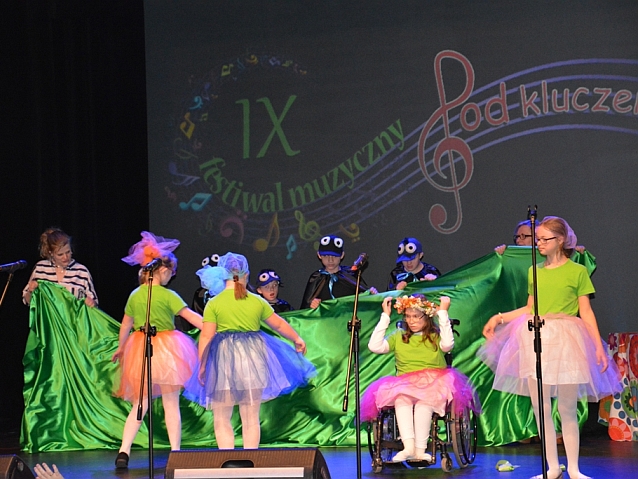 Grupa dzieci, w tym dziewczynka na wózku, na scenie w przebraniach tańczy z płachtą zielonego materiału