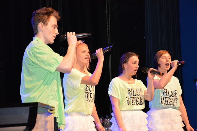 Trzy młode kobiety i młody mężczyzna śpiewają do mikrofonów na scenie