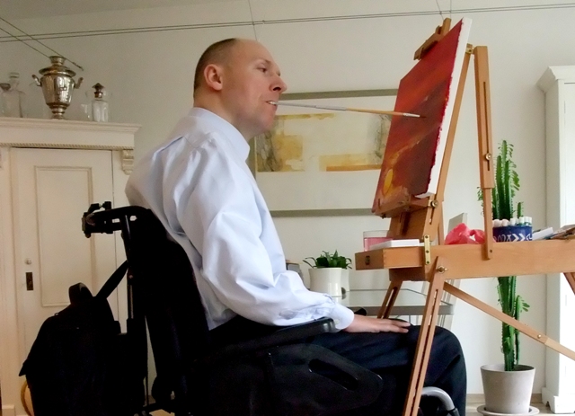 Piotr Pawłowski maluje obraz siedząc na wózku i trzymając pędzel w ustach
