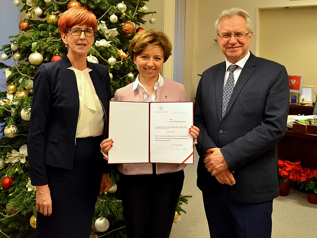 Trzy osoby pozują do zdjęcia. Od lewej: Elżbieta Rafalska, Marlena Maląg, która trzyma nominację na papierze w eleganckiej obwolucie, oraz Krzysztof Michałkiewicz