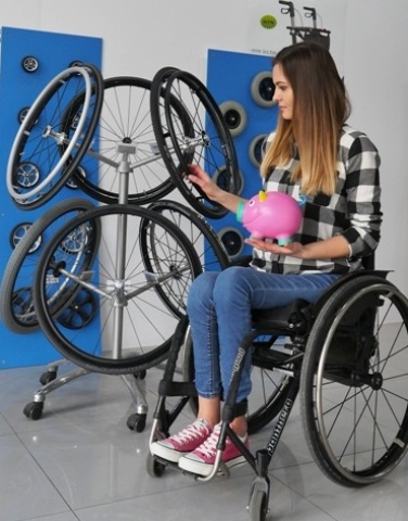 Młoda kobieta ogląda nowe koła do wózka inwalidzkiego w sklepie, a w drugiej dłoni trzyma skarbonkę