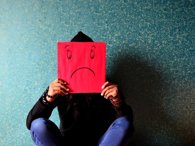 postać siedzi pod ścianą i przy twarzy trzyma kartkę, na której jest narysowana smutna mina