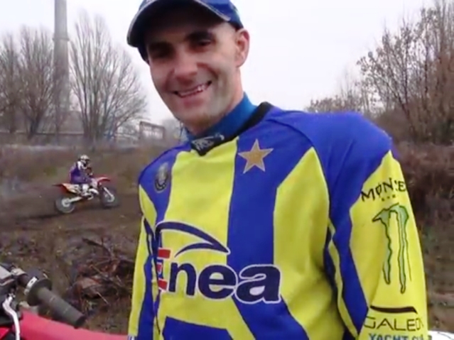 Uśmiechnięty Tomasz Gollob, w tle motocyklista mknie po torze do crossu