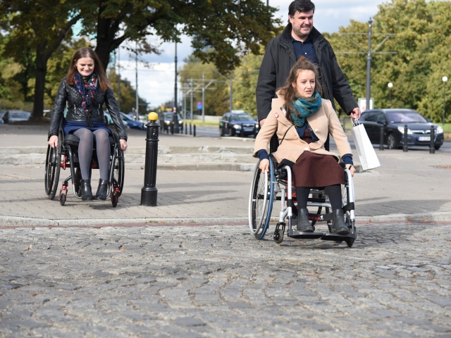 Hanna Gospodarczyk jedzie na wózku pokonując nawierzchnię z kocich łbów. Tuż za nią idzie asekurat, a z chodnika na tą nawierzchnię zjeżdża młoda kobieta, poruszająca się na wózku