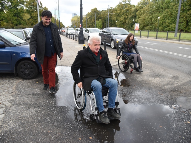 Piotr Ikonowicz przemieszcza się na wózku po dużej kałuży. Tuż za nim idzie asekurant. Po ulicy, omijając kałużę idzie młoda kobieta na wózku. Wszyscy śmieją się