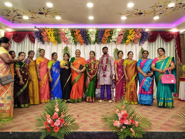 W rzędzie pozują do zdjęcia goście weselni, odświętnie i kolorowo ubrani, wśród ozdób z kwiatów