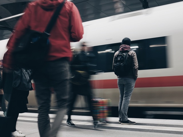 mężczyźni stoją na peronie na któym stoi pociąg, ich sylwetki są lekko zamazane