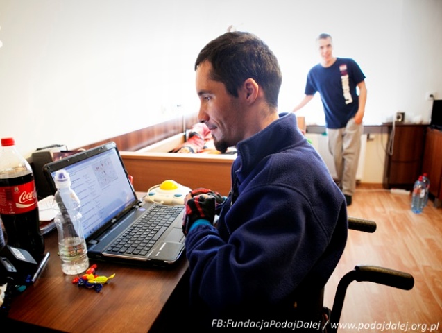 mężczyzna na wózku siedzi przed laptopem przed komputerem, w tle mężczyzna