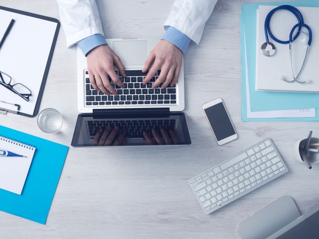 ręce lekarza w białym fartuchu na klawiaturze laptopa na biurku leży stetoskop, okulary, notatnik, termometr