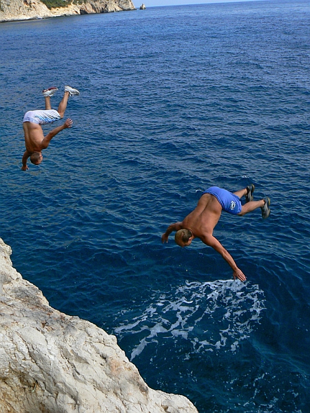 Dwóch mężczyzn w butach skacze ze skały do wody, obracając się w powietrzu