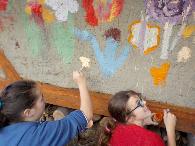 dwie uśmiechnięte dziewczynki tyłem do aparatu malują ścianę w kolorowe kwiaty