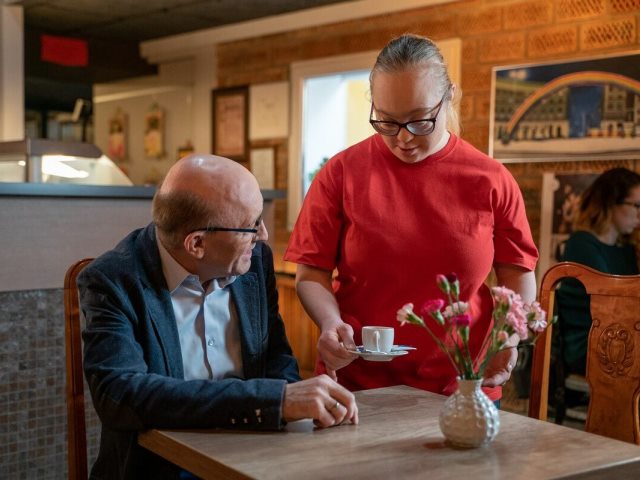 artur barciś siedzi w kawiarni przy stoliku a kawę podaje mu kobieta z zespołem downa