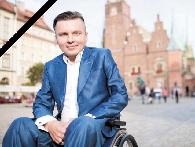 Uśmiechnięty Bartłomiej Skrzyński w niebieskim garniturze siedzi na wózku na wrocławskim rynku. Na zdjęciu jest czarna wstęga