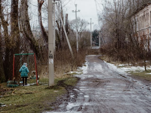 błotnista droga z resztkami śniegu po lewej drzewa wśród nich huśtawka i samotna dziewczynka po prawej zniszczona kamienica mieszkalna