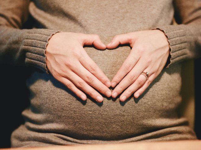 kobieta trzyma ręce na brzuchu, jest w ciąży, palce ułożone w znak serca