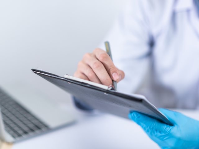 lekarz trzyma w ręku podkładkę na której pisze jedna ręka w niebieskiej rękawiczce na stole laptop