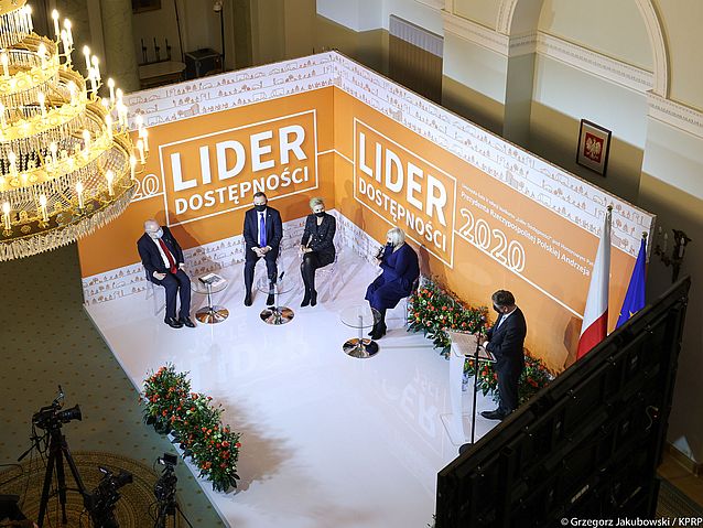 Widok z góry na scenę, na której siedzą cztery osoby, w tym: prezydent Andrzej Duda z małżonką i Ewa Pawłowska. Wokół nich bandy z napisami: Lider Dostępności 2020