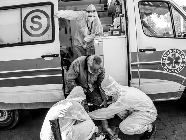 bezdomny mężczyzna siedzi przy ambulansie, obok odsłoniętej spuchniętej nogi siedzi dwóch medyków, trzeci wychyla się z wnętrza ambulansu