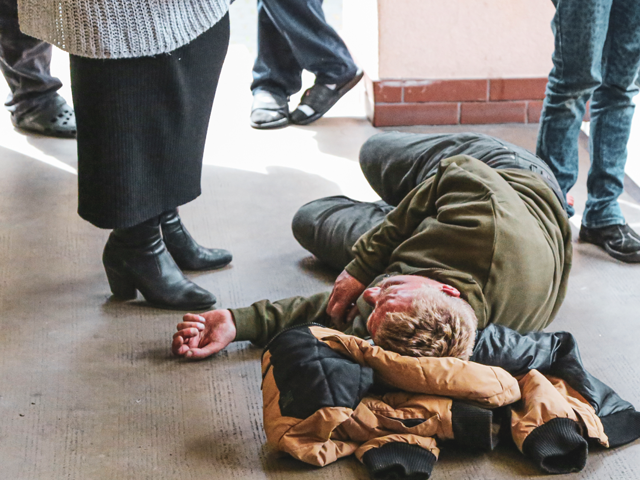 bezdomny mężczyzna leży na podłodze, ma pod głową kurtkę, obok niego widać nogi stojących nad nim lub obok ludzi