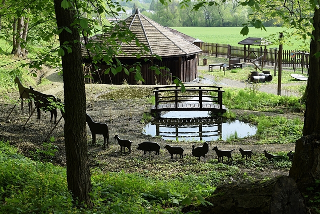 widok na okrągły drewniany dom, mostek nad małym oczkiem wodnym, rząd różnych zwierząt namalowanych na deskach