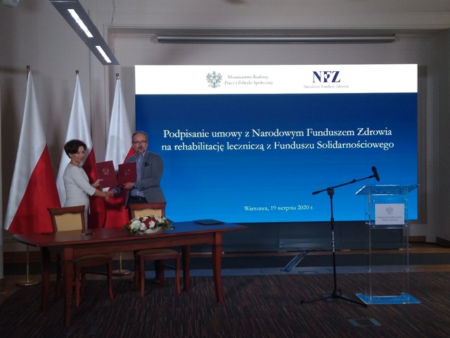 Od lewej minister marlena maląg ściska rękę Adamowi Niedzielskiemu w rękach trzymają teczkę z umową na tle trzech flag Polski