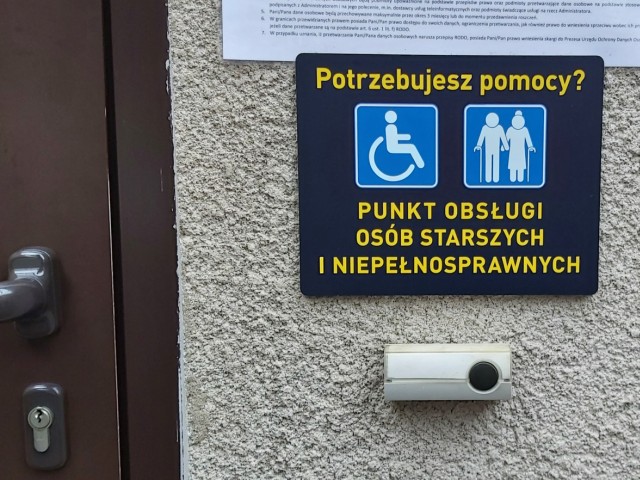zdjęcie tabliczki przy wejściu z napisem potrzebujesz pomocy pod spodem logo osoby na wózku i pary osób starszych, punkt obsługi osób starszych i niepełnosprawnych