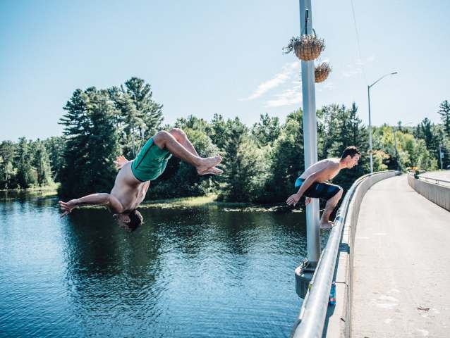 dwóch młodych mężczyzn skacze z fikołkami z mostu do rzeki