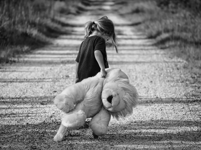 czarno białe zdjęcie z kilkuletnią dziewczynką z opuszczoną głową ciągnie za sobą dużego pluszaka idąc drogą