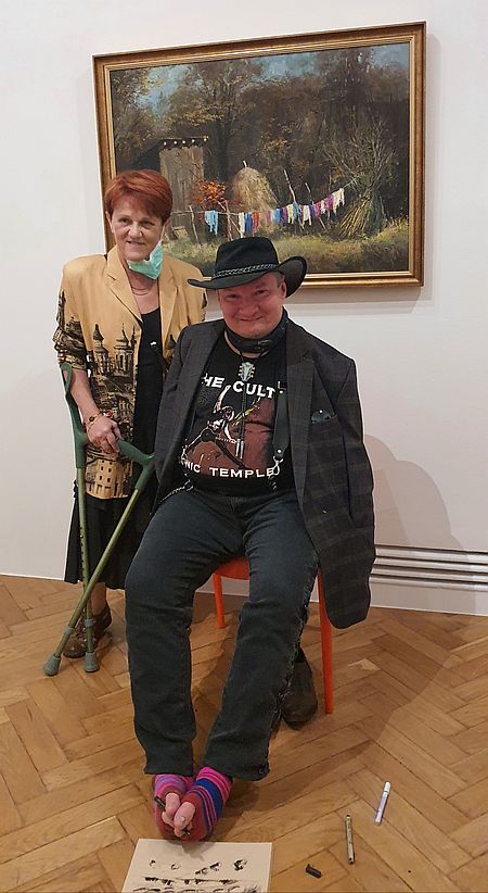 Stanisław Kmiecik maluje nogami obraz leżący na podłodze, za nim stoi kobieta o kulach, w tle obraz