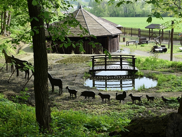 widok na skansen z góry w okręgu stoją figury różnych zwierząt i widać okrągły budynek z drewnianym dachem i małe oczko wodne