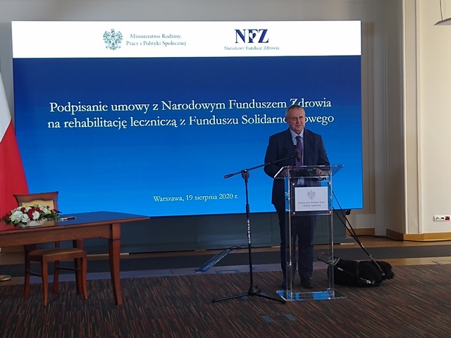 Paweł Wdówik przy mównicy na tle ekranu z napisem podpisanie umowy z narodowym funduszem Zdrowia na rehabilitację leczniczą z funduszu solidarnościowego