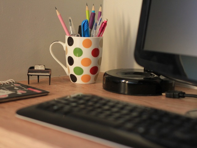 na biurku stoi kubek w kolorowe grochy z przyborami do pisania leży klawiatura notes i stoi komputer