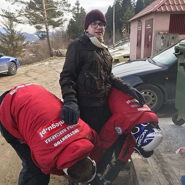 Dwie osoby zakładają buty narciarskie Wiktorowi, który uśmiechnięty patrzy w obiektyw