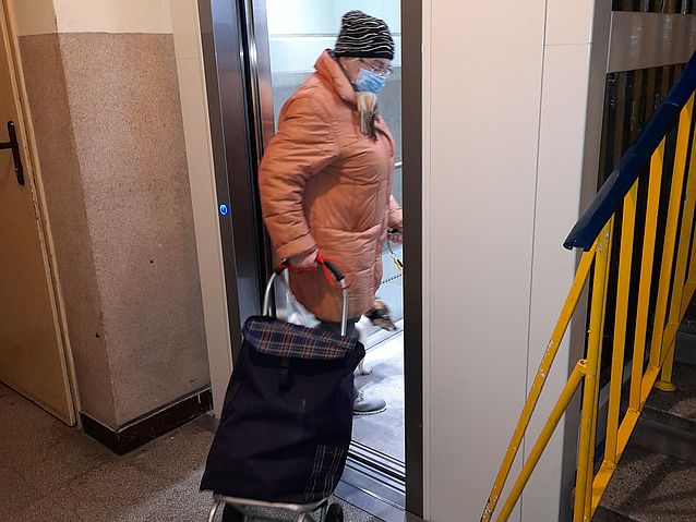 Starsza pani wchodzi do windy, ciągnąć za sobą wózek na zakupy na kółkach